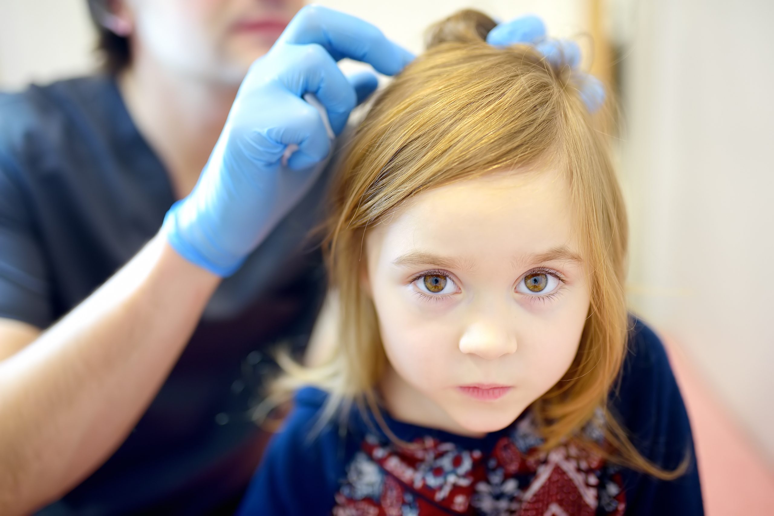 A clinician parts a young girl's hair under bright clinical lighting to examine the scalp for nits and lice during a professional screening.