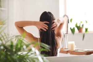 A young mother sits in a bright bathroom applying coconut oil to her own hair from a glass jar, the kind of natural treatment many parents try at home before booking a professional lice removal.