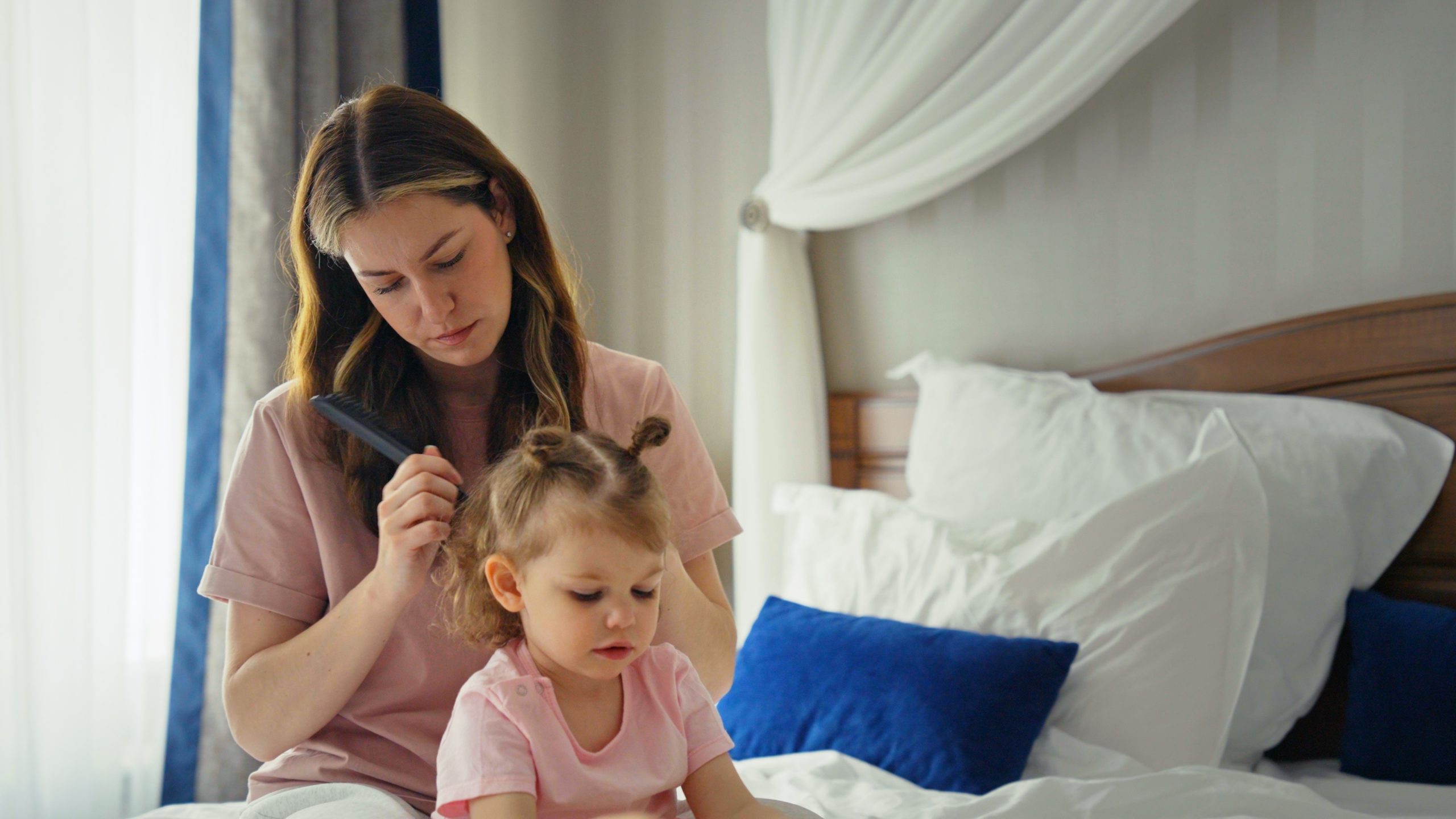 Mother and daughter bonding during a morning routine, sitting on a bed while the mom gently combs the child's hair, creating a moment of family intimacy ... See More